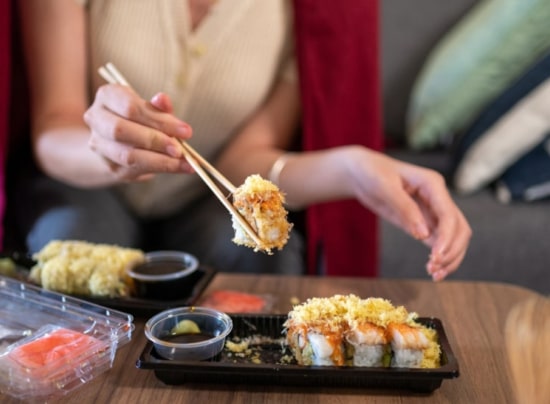 A woman holding chopsticks picks up a piece of sushi from a takeout container.